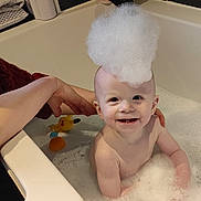Erik is registered to the contest to win money with this photo: baby, infant, bathtub, bubbles, foam, bubble_hat, smile, happy, bath_toys, pikachu_toy, adult_hand, water, tile_wall, bathroom, face, teeth, skin, tub_rim, sitting, playful