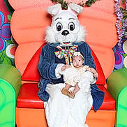 Aylin is registered to the contest to win money with this photo: baby, child, easter_bunny, costume, chair, colorful, flower, headband, indoor, person, plush, seat, smiling, toy, whimsical, decor, green, orange, red, blue