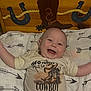baby, smiling_baby, onesie, cowboy, wooden_chest, horseshoe, boot_graphic, arrow_pattern, bedding, nursery, happy, face, head, lying_down, infant, fabric, decor, text_on_clothing, portrait, cute