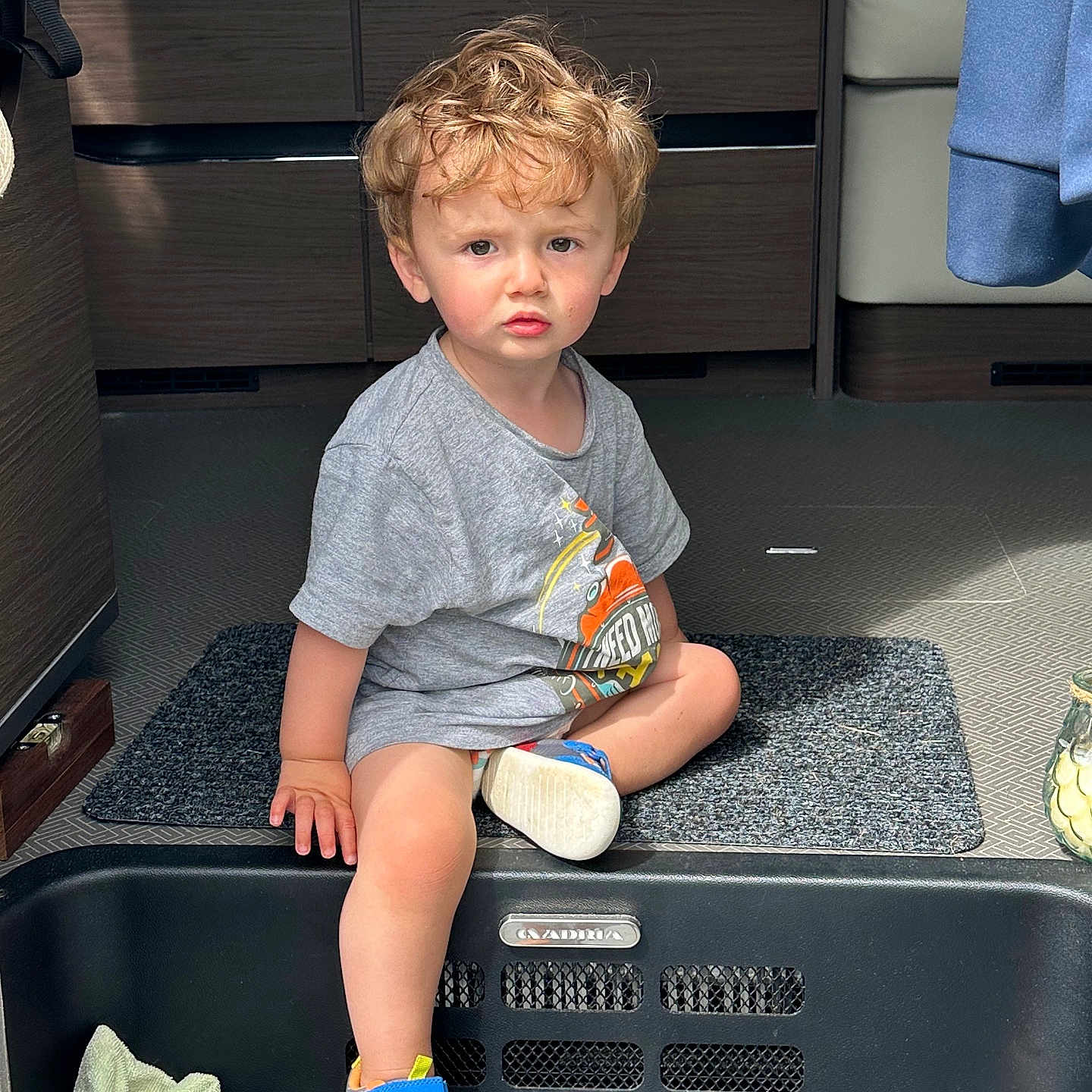 Hew is registered to the contest to win money with this photo: carpet, casual_clothing, child, contemplative, curly_hair, daylight, expression, floor_mat, furniture, grey_tshirt, hand, indoor, modern_interior, person, shoe, sitting, sneakers, step, toddler, young_child