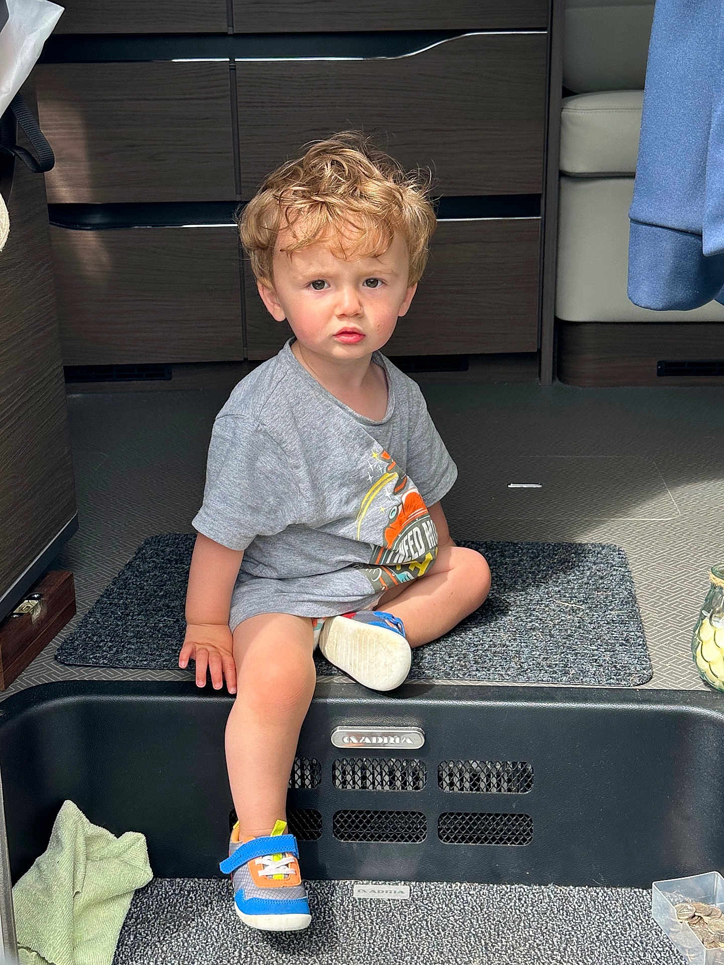 Hew is registered to the contest to win money with this photo: toddler, child, indoor, sitting, curly_hair, grey_tshirt, sneakers, step, floor_mat, modern_interior, carpet, expression, young_child, casual_clothing, person, daylight, contemplative, shoe, hand, furniture