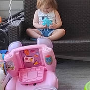 Robin joined the competition — help win amazing prizes! baby_toy, blue_toy, casual_clothing, child, curly_hair, daylight, fence, floor, outdoor, pink_toy, plastic_toy, playtime, porch, relaxing, sitting, small_toys, toddler, toy, white_lattice, wicker_swing