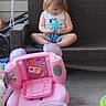 baby_toy, blue_toy, casual_clothing, child, curly_hair, daylight, fence, floor, outdoor, pink_toy, plastic_toy, playtime, porch, relaxing, sitting, small_toys, toddler, toy, white_lattice, wicker_swing