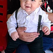 Parker joined the competition — help win amazing prizes! baby, child, person, suspenders, white_shirt, black_pants, barefoot, lap, hand, red_chair, casual_clothing, indoor, curious, smiling, seated, cute, portrait, young_child, adult_hand, jeans