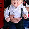 baby, child, person, suspenders, white_shirt, black_pants, barefoot, lap, hand, red_chair, casual_clothing, indoor, curious, smiling, seated, cute, portrait, young_child, adult_hand, jeans