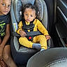 child, toddler, baby, siblings, car, car_seat, seatbelt, safety_harness, backseat, vehicle, portrait, indoor, sitting, yellow_outfit, black_shirt, curly_hair, smiling, looking_at_camera, family, safe_travel