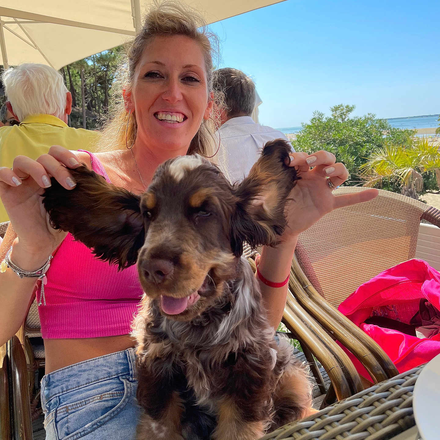 Pascal Ravel participe au concours pour gagner de l'argent avec cette photo : woman, dog, smiling, outdoor, cafe, sunny, blue_sky, greenery, casual_clothing, floppy_ears, happy, sitting, table, chair, umbrella, bracelet, jeans, pink_top, portrait, playful