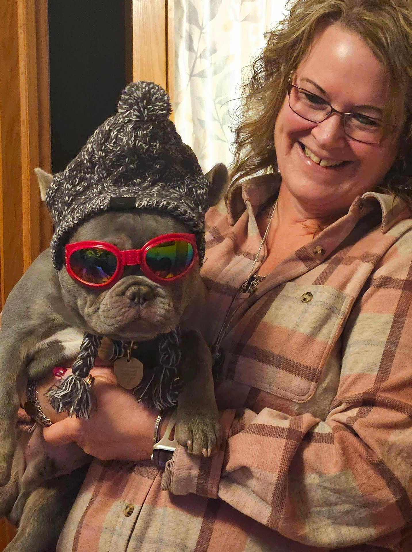 Thumper is registered to the contest to win money with this photo: accessory, curtain, dog, dog_face, eyeglasses, french_bulldog, holding, human_face, indoor, knit_hat, necklace, paw, pet, plaid_shirt, pom_pom_hat, portrait, smile, sunglasses, woman, wooden_door
