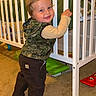 Kollins joined the competition — help win amazing prizes! baby, barefoot, camo_vest, carpet, child, crib, cute, face, furniture, hair, hand, indoor, pants, person, playroom, room, smile, standing, toddler, toy