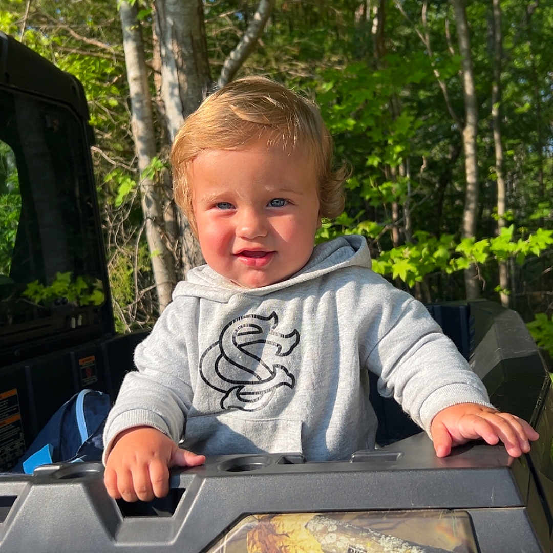 Kollins joined the competition — help win amazing prizes! blonde_hair, blue_eyes, camouflage, casual_clothing, child, curious, daylight, forest, grip, hoodie, leaves, nature, outdoor, person, playful, sunlight, toddler, trees, vehicle, young_child