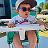 toddler, child, sunglasses, baseball_cap, high_chair, outdoor, sunny, green_chair, table, footwear, shorts, white_tshirt, baby, person, summer, shade, recreational_vehicle, bicycle, grass, daytime