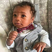 D'Vaun is registered to the contest to win money with this photo: baby, child, infant, vest, bow_tie, clothing, curly_hair, patterned_background, hand, face, eyes, portrait, cute, indoors, sitting, expression, fashion, person, young, adorable