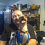 man, dog, smiling, happy, indoor, kitchen, table, water_bottle, sitting, clothing, blanket, refrigerator, shelf, container, plastic_bag, pot, collar, tag, tongue, facial_hair