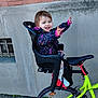 child, toddler, person, bicycle, bike, child_seat, wheel, saddle, colorful_jacket, smiling, happy, outdoor, brick_wall, concrete_wall, pavement, shoe, legs, handlebar, playful, seat