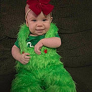 Izabella is registered to the contest to win money with this photo: baby, child, clothing, costume, couch, cute, face, furniture, fuzzy, green_fur, happy, headband, indoor, infant, person, portrait, red_bow, sitting, smile, young