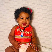 Hannah joined the competition — help win amazing prizes! baby, child, smiling, curly_hair, red_bows, orange_top, sitting, blanket, happy, cute, infant, portrait, skin, barefoot, necklace, indoors, plush, person, toddler, comfortable