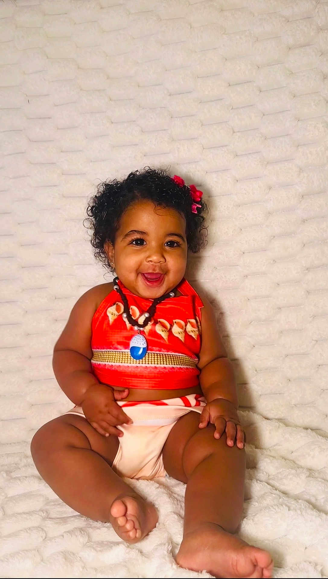Hannah joined the competition — help win amazing prizes! baby, child, smiling, curly_hair, red_bows, orange_top, sitting, blanket, happy, cute, infant, portrait, skin, barefoot, necklace, indoors, plush, person, toddler, comfortable