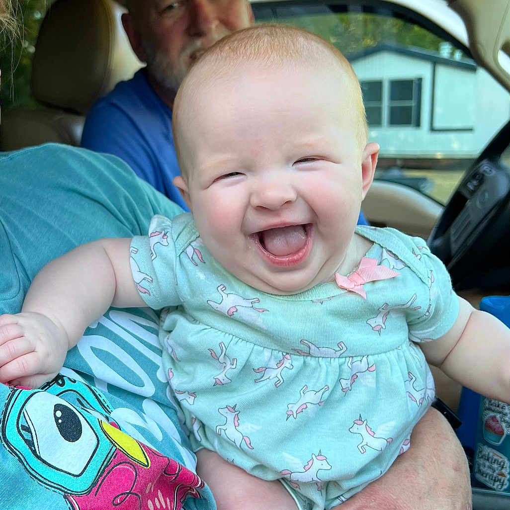 Bexlee is registered to the contest to win money with this photo: baby, child, infant, smiling, laughing, portrait, car, vehicle, indoors, adult, caregiver, family, onesie, clothing, unicorn_pattern, hands, seated, happy, close_up, daylight