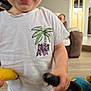 toddler, child, white_tshirt, banana, living_room, toy, couch, person, floor, smiling, indoor, furniture, hand, blur, casual_clothing, young_child, happy, palm_tree_design, holding, background
