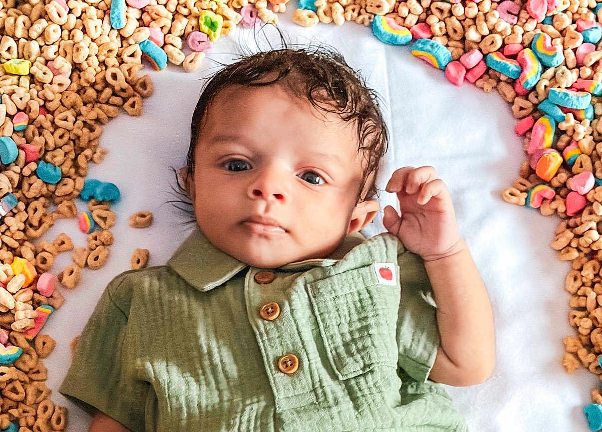 Davion is registered to the contest to win money with this photo: baby, infant, child, newborn, portrait, face, eyes, hand, green_shirt, buttons, lying_down, white_blanket, breakfast_cereal, cereal_loops, colorful_marshmallows, cute, soft_texture, closeup, hair, playful_setup