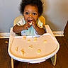 toddler, child, high_chair, food, bib, eating, mouth_open, curly_hair, indoor, wooden_floor, messy, plate, snack, small_child, cute, person, baby, feeding, happy, young