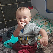 Ezekiel joined the competition — help win amazing prizes! toddler, child, baby, indoor, bed, pillow, clothing, grey_shirt, red_pants, face, hand, playing, toy, curious, expression, home, blanket, wall, floor_tiles, person
