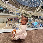 Julie-Victoire participe au concours pour gagner de l'argent avec cette photo : child, toddler, indoor, mall, shopping_mall, plush_toy, fox_toy, white_sweater, curly_hair, floor, glass_railing, chandelier, modern_architecture, people, shopping, escalator, lighting, curious, crouching, fashion