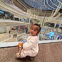 child, toddler, indoor, mall, shopping_mall, plush_toy, fox_toy, white_sweater, curly_hair, floor, glass_railing, chandelier, modern_architecture, people, shopping, escalator, lighting, curious, crouching, fashion