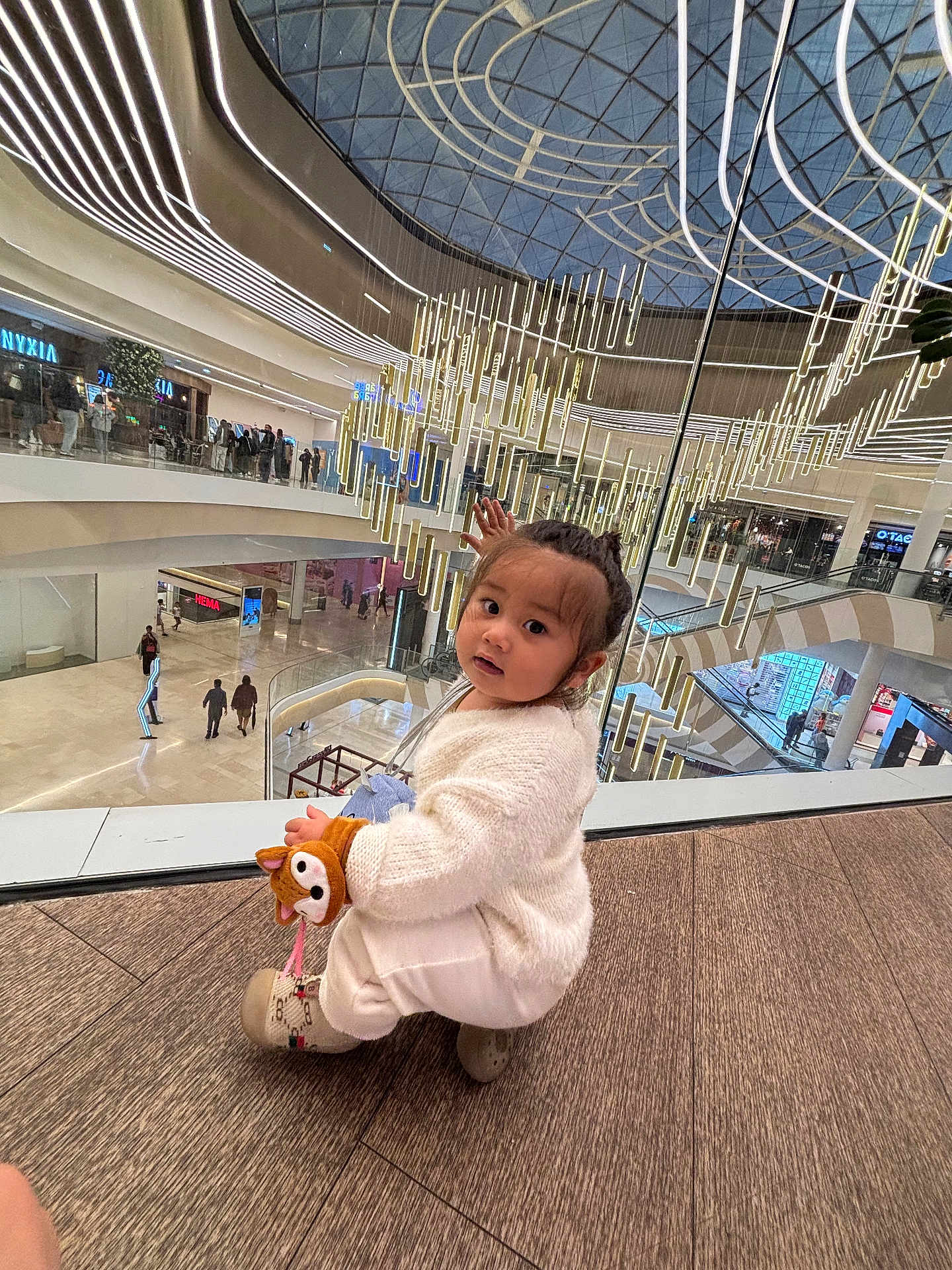 Julie-Victoire participe au concours pour gagner de l'argent avec cette photo : child, toddler, indoor, mall, shopping_mall, plush_toy, fox_toy, white_sweater, curly_hair, floor, glass_railing, chandelier, modern_architecture, people, shopping, escalator, lighting, curious, crouching, fashion