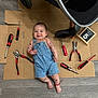 baby, denim_overalls, tools, cardboard, smiling, infant, floor, stroller, pliers, screwdrivers, chalkboard, milestone, red_bracelet, black_marks, footwear, wooden_floor, person, happy, indoor, playful