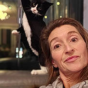 Tickle is registered to the contest to win money with this photo: woman, cat, black_and_white_cat, hoodie, indoor, reflection, stretching_cat, face, brown_hair, casual_clothing, expression, closeup, portrait, furniture, blurred_background, light, window, home, relaxed, playful