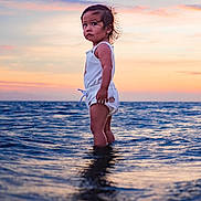 Tui is registered to the contest to win money with this photo: toddler, child, ocean, water, sunset, sky, waves, standing, portrait, outdoor, nature, sea, reflection, clothing, person, water_body, sunlight, young_child, explorer, adventure