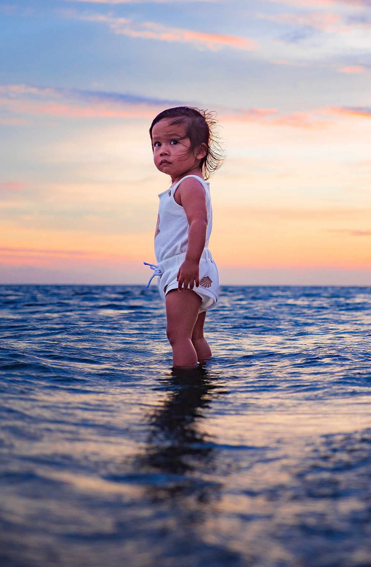 Tui is registered to the contest to win money with this photo: toddler, child, ocean, water, sunset, sky, waves, standing, portrait, outdoor, nature, sea, reflection, clothing, person, water_body, sunlight, young_child, explorer, adventure
