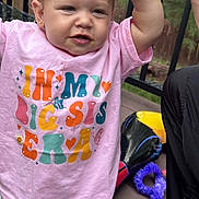 Elliana joined the competition — help win amazing prizes! toddler, child, pink_shirt, colorful_text, outdoor, deck, toys, playful_expression, raising_arm, casual_clothing, earring, curly_hair, person, summer, daylight, wooden_rail, purple_toy, black_pants, patterned_pants, cute