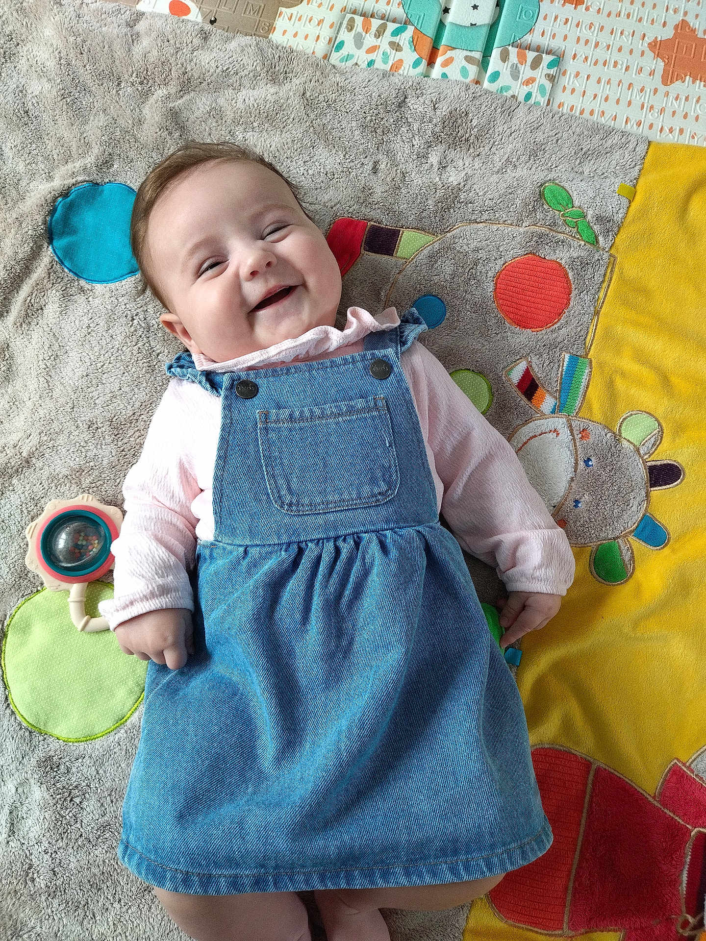 Lyanna a rejoint le concours — aidez-le/la à gagner de superbes lots ! baby, smiling, denim_dress, pink_shirt, play_mat, toy, happy, child, infant, lying_down, colorful, soft_texture, cute, indoor, portrait, person, clothing, face, baby_toy, playful
