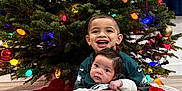 Milani is registered to the contest to win money with this photo: baby, blanket, candid, child, christmas_tree, clothing, decorations, family, floor, happy, holiday, indoor, lights, ornaments, portrait, sitting, smile, sock, toddler, together