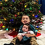 Milani is registered to the contest to win money with this photo: child, baby, christmas_tree, holiday, decorations, ornaments, lights, sitting, blanket, smile, happy, indoor, clothing, sock, floor, candid, family, toddler, portrait, together