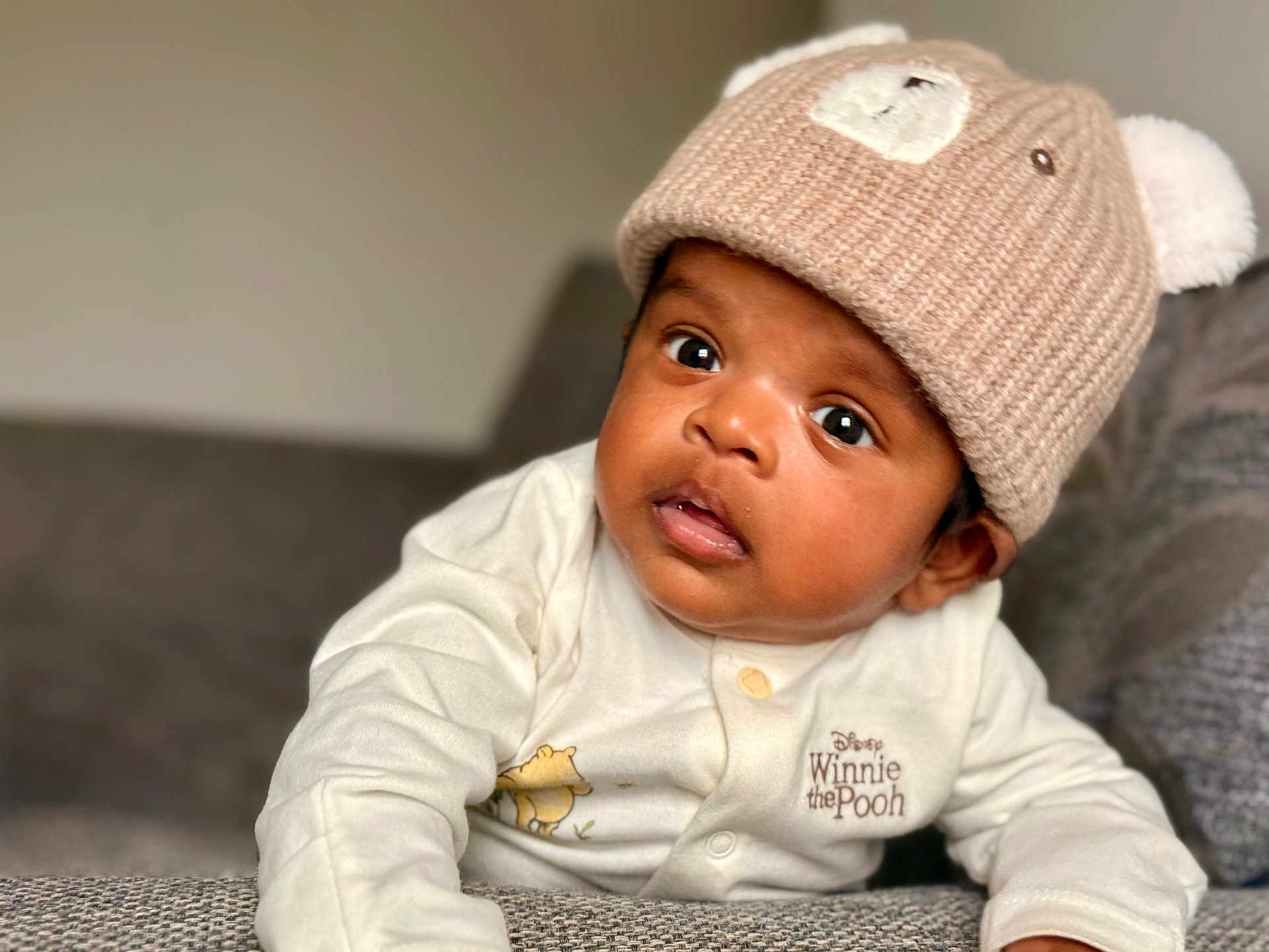 Aadhav joined the competition — help win amazing prizes! baby, infant, child, knitted_hat, bear_hat, winnie_the_pooh, onesie, clothing, face, eyes, mouth, couch, furniture, indoor, portrait, cute, curious, expression, young_child, soft_lighting