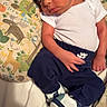 Kingston is registered to the contest to win money with this photo: baby, infant, hat, sneakers, pants, white_shirt, pillow, animal_print, bed, curious_expression, child, clothing, footwear, person, indoors, cute, newborn, resting, colorful, soft_lighting