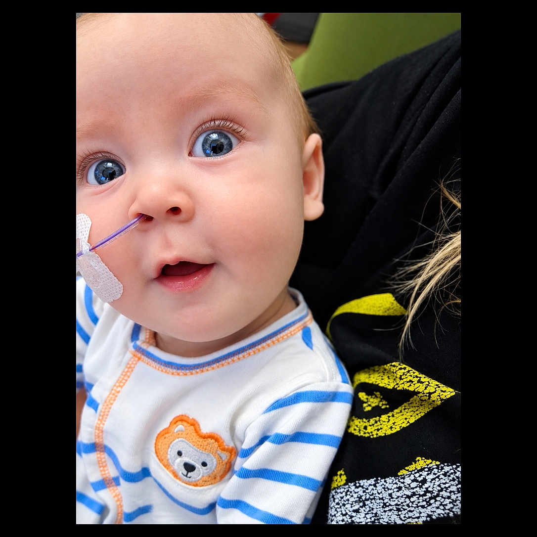 Ryder is registered to the contest to win money with this photo: baby, infant, child, portrait, close_up, face, eyes, wide_eyed, smiling, indoor, medical_tube, tape, healthcare, striped_clothing, onesie, teddy_bear, caregiver, black_clothing, green_background, photography