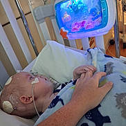Ryder is registered to the contest to win money with this photo: baby, hospital, crib, medical_patches, nasal_tube, hand_holding, blanket, toy, blue_light, child, bed, healthcare, infant, person, medical_equipment, comfort, care, indoor, closeup, tender_moment