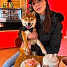 dog, shiba_inu, woman, person, portrait, smiling, cafe, indoors, bow_tie, black_jacket, baseball_cap, long_hair, cupcake, tea, teapot, table, food, colorful_background, pet, sitting