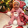 baby, child, christmas_tree, christmas_lights, holiday, pajamas, santa_hat, smiling, gift_wrap, decorations, floor, indoors, festive, seasonal, happy, cute, person, celebration, red_and_white, christmas_stocking