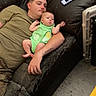 baby, infant, child, adult, parent, father, couch, sitting, lying_down, green_outfit, home, living_room, portrait, face, two_people, relaxed, game_controller, snacks, baby_gate, indoor