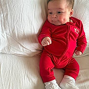 Reuben is registered to the contest to win money with this photo: baby, infant, red_onesie, white_sneakers, dimples, bed, white_bedspread, soft_texture, cute, child, portrait, relaxed, indoors, young_child, footwear, sleepwear, expression, skin, person, small_hand