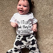 Renley is registered to the contest to win money with this photo: baby, child, smiling, headband, white_shirt, printed_text, cow_print_pants, carpet, lying_down, happy, cute, infant, person, indoors, playful, fashion, clothing, portrait, funny_text, closeup