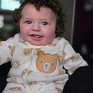 Lilith is registered to the contest to win money with this photo: baby, child, curly_hair, onesie, teddy_bear_pattern, smiling, tongue_out, indoor, portrait, cute, happy, infant, soft_fabric, cozy, young_child, face, person, sitting, warm_clothing, playful