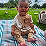baby, child, smiling, sitting, blanket, park, grass, outdoor, bib, pants, lion_design, footwear, people, trees, sky, clouds, bag, playground, daytime, cute