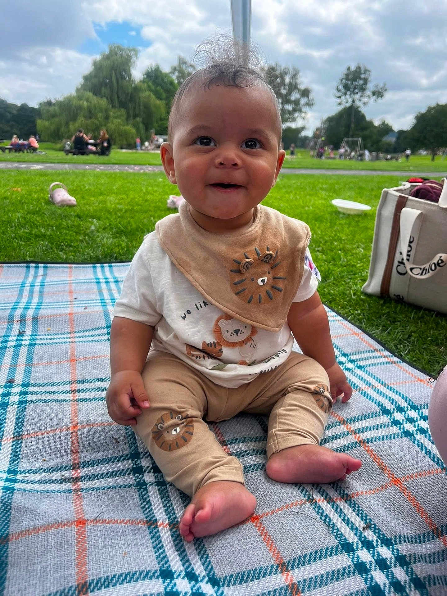 Ezra joined the competition — help win amazing prizes! baby, child, smiling, sitting, blanket, park, grass, outdoor, bib, pants, lion_design, footwear, people, trees, sky, clouds, bag, playground, daytime, cute