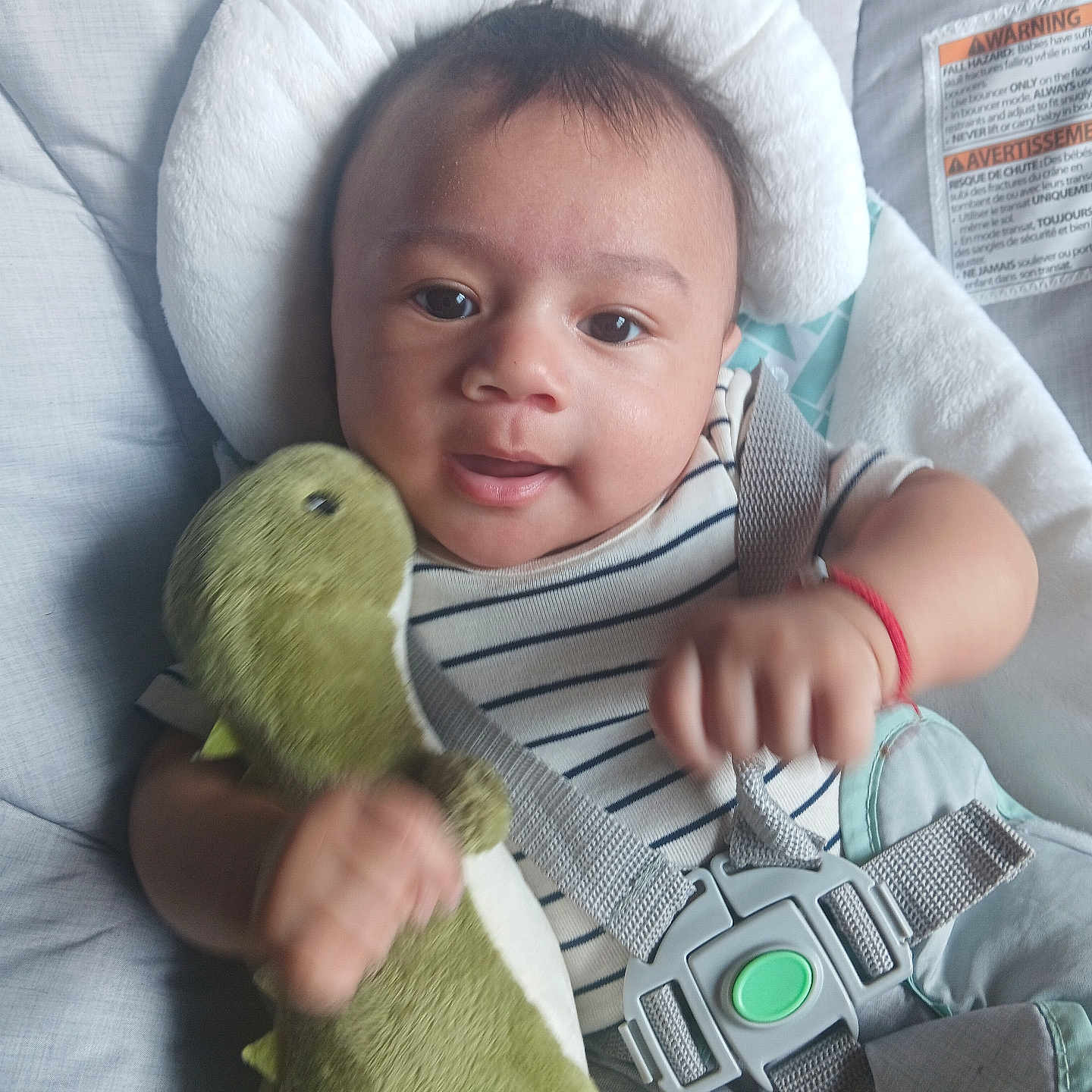 Julian is registered to the contest to win money with this photo: baby, infant, child, plush_toy, dinosaur_toy, car_seat, safety_harness, striped_shirt, smiling, happy, cute, indoors, cushion, headrest, face, person, young_child, red_bracelet, soft_toy, closeup
